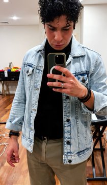 A young man with curly hair taking a mirror selfie, wearing a black shirt under a light blue denim jacket and olive green pants, with a ring on his finger.
