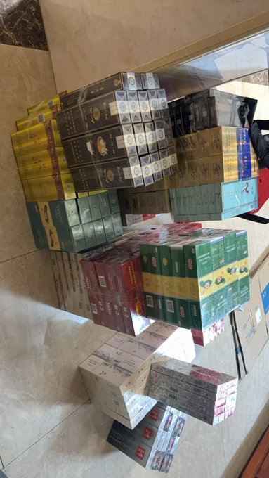 Stacks of various boxed products piled up in a warehouse or store room, including what appears to be tea or oil cartons in green, gold, and red packaging.
