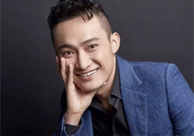 A smiling man in a blue blazer and black shirt rests his chin on his hand against a dark backdrop, embodying the confident, ambitious spirit celebrated in the birthday tribute to Justin Sun.