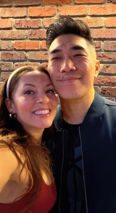 A close-up selfie of a smiling woman with long brown hair wearing a red top and a headband, posing next to an Asian man with styled black hair wearing a black jacket, with an exposed brick wall in the background.