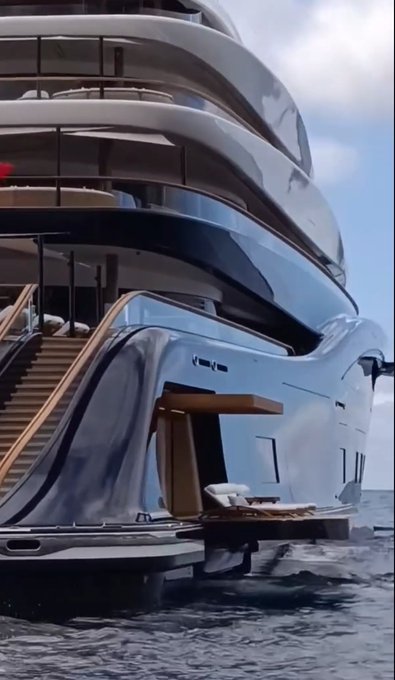 The image features a large yacht with multiple decks, including one that has stairs leading up to it. The yacht appears to be floating on water, possibly in the ocean or a lake. The surrounding area seems to have some vegetation, as there are trees visible near the yacht.