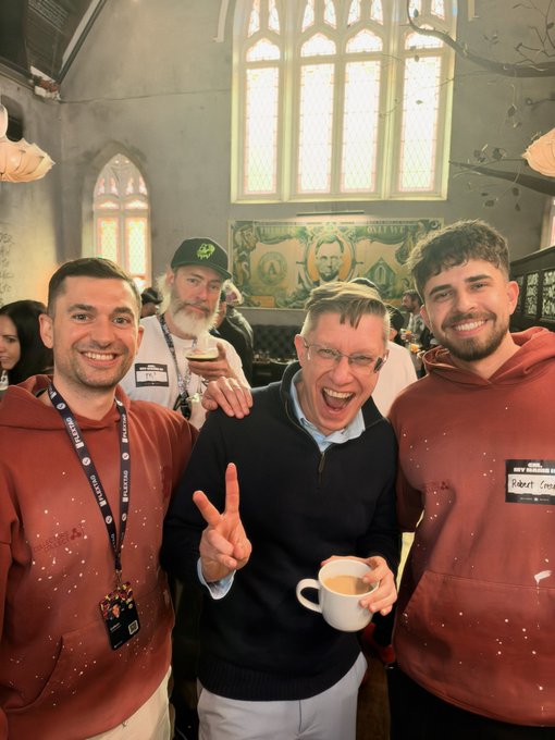 Four men posing cheerfully inside what appears to be a church-like venue with arched windows; two wear maroon hoodies, one wears a black sweater and peace-signs while holding a mug, and another with a white beard wears a black cap.