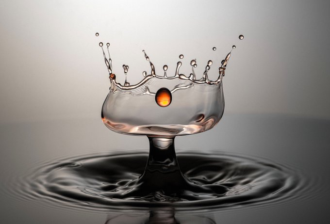 A crown-shaped water splash frozen in time with a glowing amber orb at its center, rising from concentric ripples below. This macro splash photograph captures the ephemeral beauty of liquid art, perfectly embodying the "Amber Crown 2" NFT's concept of a crown that forms, breaks, and vanishes in an instant.