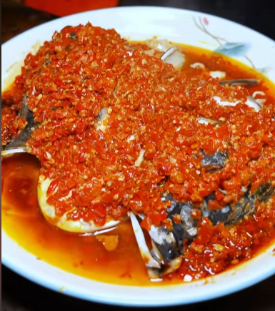 A close-up photo of a traditional Chinese dish featuring a steamed fish head covered in a generous mound of chopped red chili peppers sitting in oily red chili sauce on a white plate.