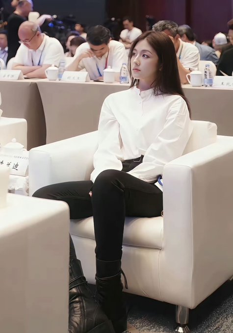 A young woman with long brown hair, wearing a white blouse and black pants with boots, sits in a white armchair at what appears to be a formal conference or panel event with attendees in the background.