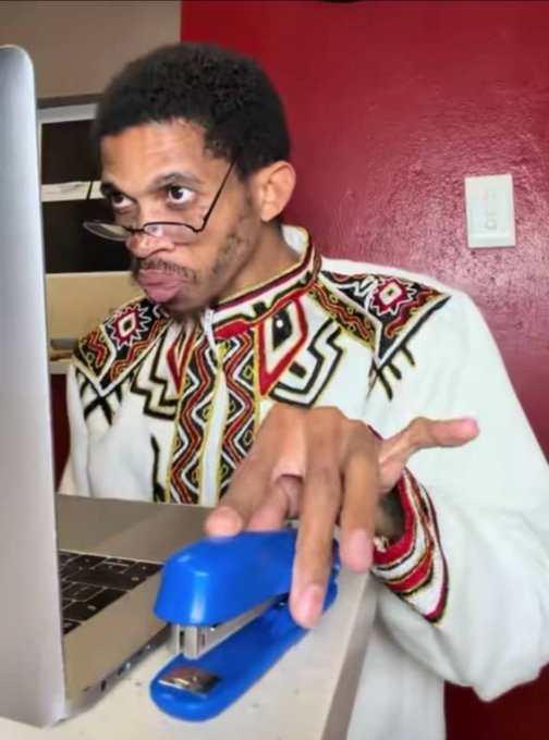 A man wearing glasses and a patterned dashiki shirt is operating a blue stapler on a desk next to a laptop in what appears to be a humorous meme-style image.