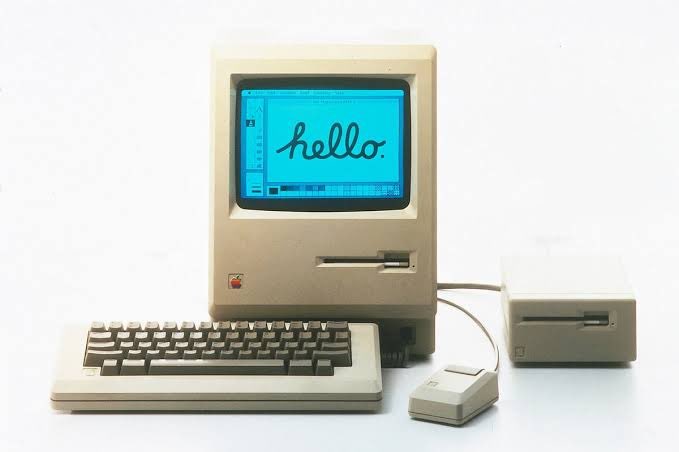 A vintage Apple Macintosh computer setup with keyboard, mouse, and external drive, displaying the word 'hello.' in cursive on a blue screen.