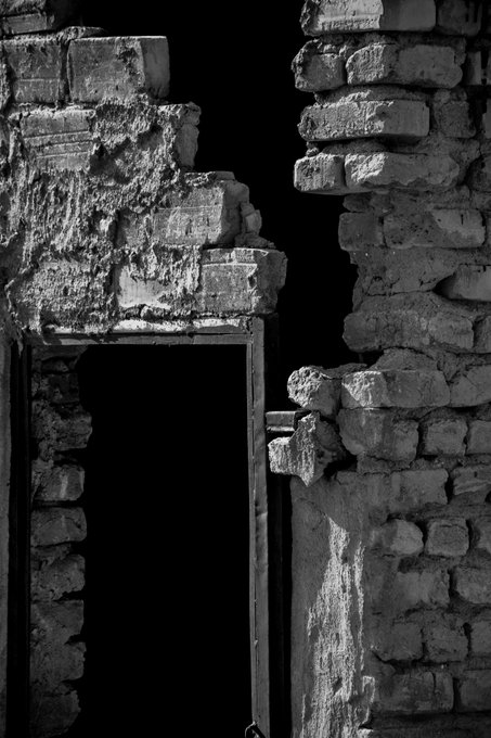 A crumbling brick wall splits open in stark monochrome, revealing an empty doorway swallowed by absolute darkness. The image echoes the caption's poetic imagery of collapse and shadow, embodying the unsettling quiet of a structure fractured by time—much like the haunting, minimalist world of the referenced *Cleft*.