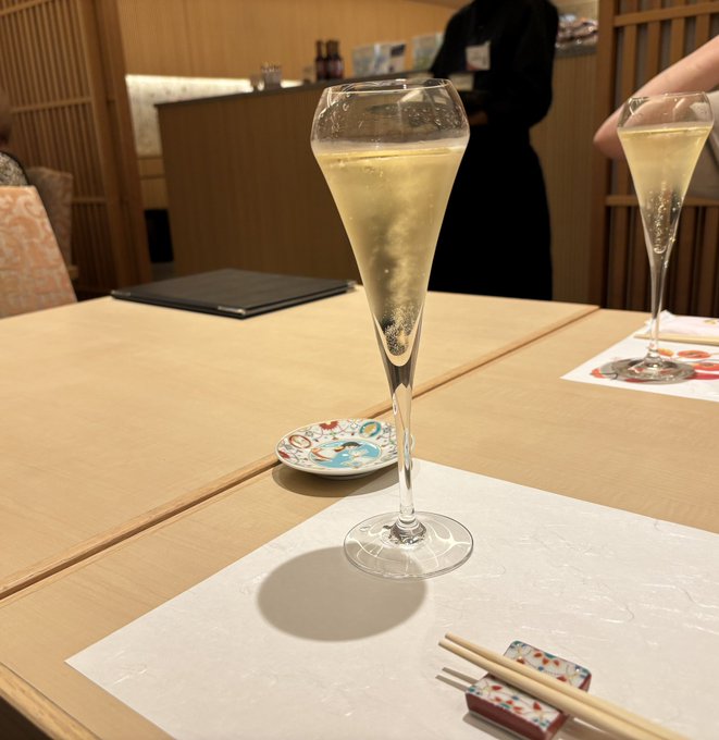 A champagne flute filled with sparkling wine sits on a white placemat on a wooden table, with a smaller dish and chopsticks nearby.
