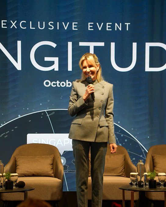 Yana Prikhodchenko, CEO of Cointelegraph, addresses the audience at LONGITUDE's 10th edition exclusive event in Singapore in October, holding a microphone in front of a branded backdrop while standing on a panel stage with chairs and small tables nearby.