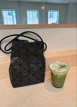 A sleek black geometric-pattern handbag sits next to a green iced matcha drink on a wooden café counter, perfectly capturing the post's "steady progress and good matcha" mantra as a stylish visual reminder to start the week intentionally.