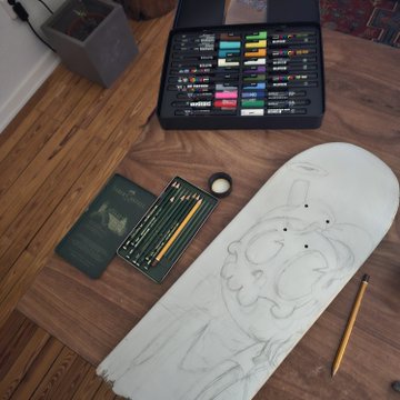 An overhead view of an art workspace featuring a skateboard deck with a pencil sketch, an open set of colored markers, pencils, and a passport on a wooden floor.