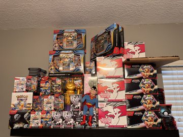 A large collection of Pokemon trading card game products, including booster boxes and tins, stacked together with a Pokemon figurine placed in front of the display.