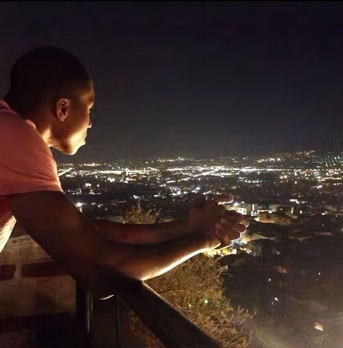 A man stands silhouetted at a railing overlooking a sprawling, illuminated cityscape at night, his hands clasped as he gazes out at the view. The contemplative scene of someone pausing to take in the vast urban landscape mirrors the mood of feeling "completely sidelined" and reflective, perhaps watching the world pass by while sidelined from the Robinhood "cat coin" action.