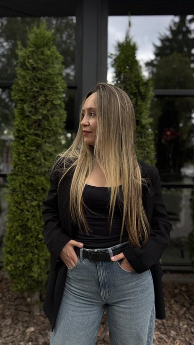 A portrait of a woman with long blonde-highlighted hair wearing a black top and blazer with blue jeans, standing outdoors in front of green shrubs.