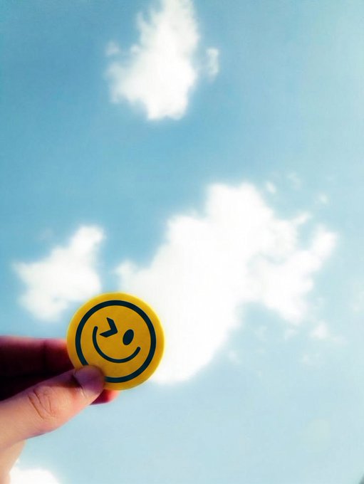 A hand holds up a yellow winking smiley face emoji sticker against a bright blue sky with fluffy white clouds, playfully responding to the prompt to describe your portfolio with a single emoji.