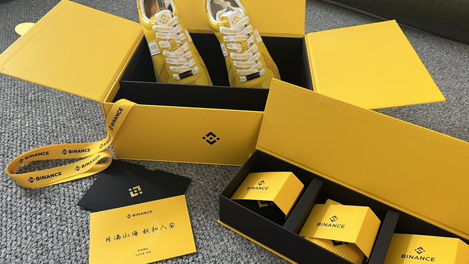 An open yellow Binance branded box containing a pair of yellow high-top sneakers, with various Binance-branded accessories like a lanyard and packaging materials on a gray surface.