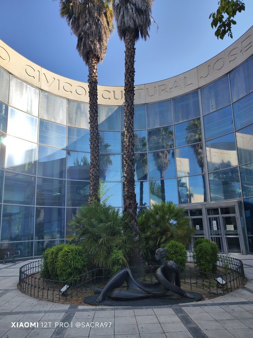 A modern glass building with curved architecture displays the text 'CIVICO CULTURALE JOSEF' along its upper edge, featuring palm trees and a reclining dark figure sculpture in front, watermarked 'XIAOMI 12T PRO | @SACRA97'.