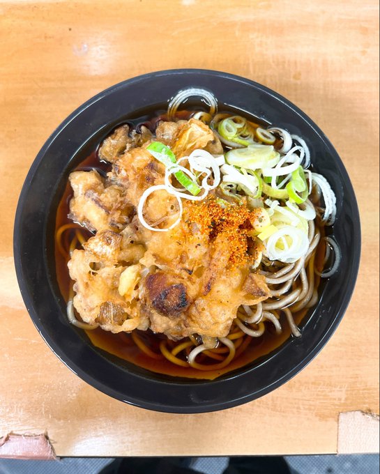 A bowl of Japanese soba noodles topped with fried meat (likely chicken or pork), green onions, and red chili spice, served in a dark broth on a wooden table.