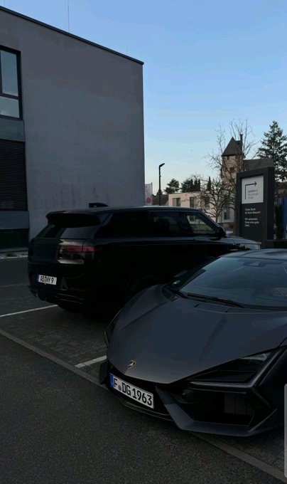 Two luxury SUVs parked outside a modern building, including a black Range Rover and a grey Lamborghini Urus with German license plates 'F DG 1963'.