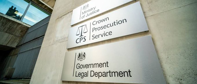 A photograph of signage outside a UK government building displays the names of three official entities: the "Ministry of Justice," the "Crown Prosecution Service (CPS)," and the "Government Legal Department." The image is used to illustrate a news story about the Crown Prosecution Service securing a £1.8M confiscation order against former NCA officer Paul Chowles for stealing bitcoin.
