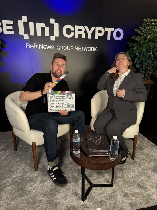 Two people sitting in chairs during an interview for 'BeInCrypto', with a man holding a clapperboard and a woman speaking into a microphone.