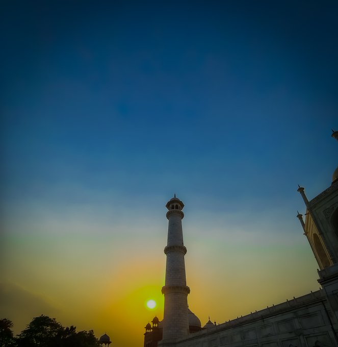 A silhouette of the Taj Mahal's towering minaret stands tall against a dramatic sunset sky, with the glowing sun dipping below in vibrant amber and blue hues. The image perfectly captures the post's caption of "Sunset Serenity," showcasing the iconic monument's serene beauty as twilight descends.