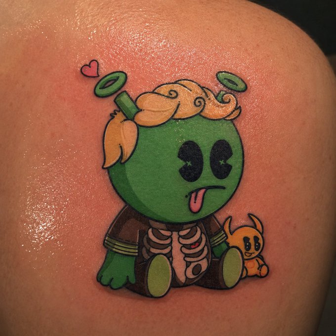 A colorful tattoo on skin depicting a green, cartoonish character with blonde hair, two halo-like rings, and a small orange creature beside it, with the green character sticking out its tongue.