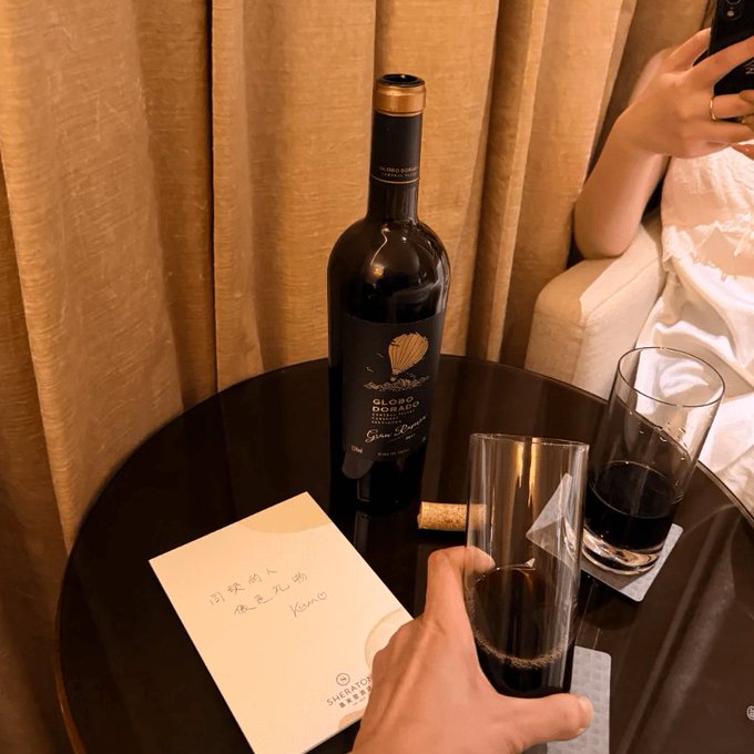 A photograph of a bottle of 'Gloria Ferrer Sonoma Brut' wine and two glasses of wine on a dark table, with someone's hand holding one glass and a handwritten note on a Sheraton-branded card.
