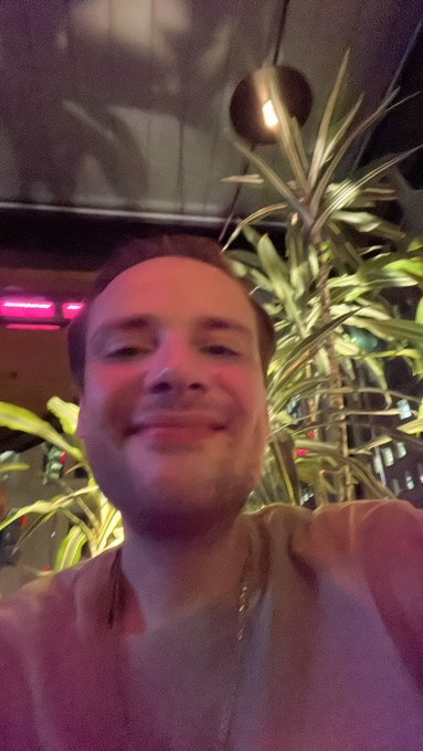 A low-angle selfie of a smiling man in a light-colored shirt, with green plants and a hanging bulb light visible in the warm-lit background.