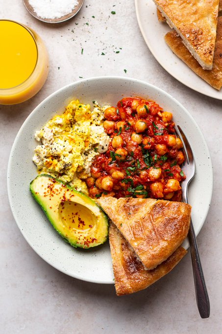 A breakfast plate featuring scrambled eggs, saucy chickpeas in tomato sauce, avocado slices, and toasted bread triangles, accompanied by a glass of orange juice.