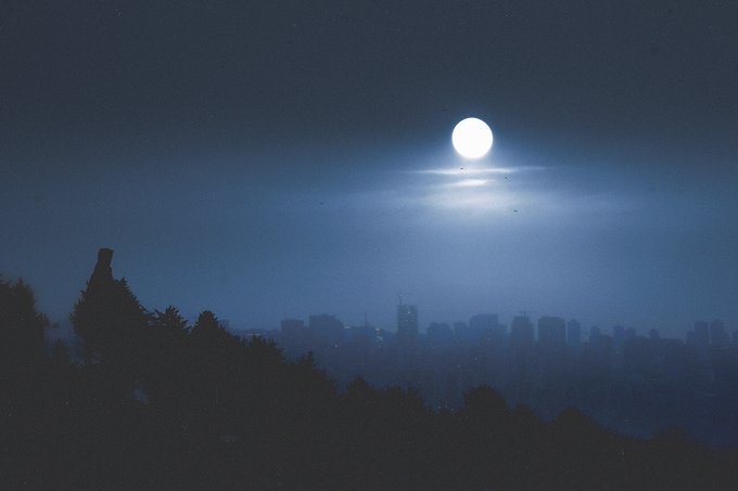 A nighttime cityscape silhouette featuring a bright full moon glowing in a deep blue sky above a hazy urban skyline and dark treeline.