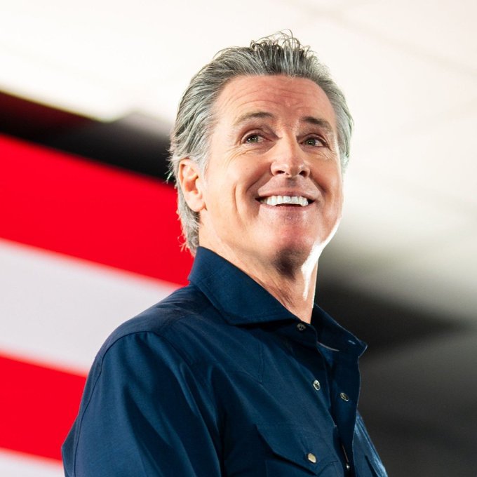 A photo of a smiling older man with gray hair wearing a dark navy shirt, standing in front of a red, white, and black striped background that resembles an American flag.