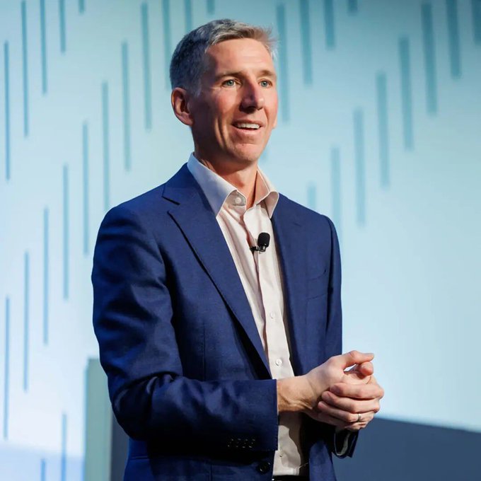 The image shows Bitwise CIO Matt Hougan speaking at what appears to be a conference, dressed in a navy blue suit with a lapel microphone, visually representing the expert quoted in the post about Trump's meeting with crypto executives being positive for Bitcoin and DeFi applications.