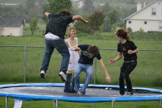 Four young people jumping and playing energetically on a blue trampoline in a grassy backyard with houses in the background.