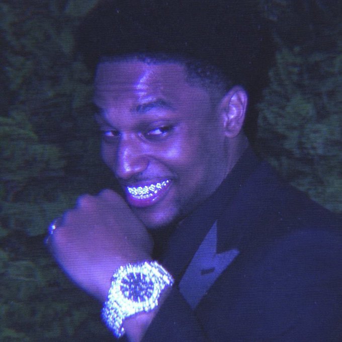 A man with diamond-encrusted grillz and a matching large diamond watch smiles at the camera in dim, purple-lit lighting.