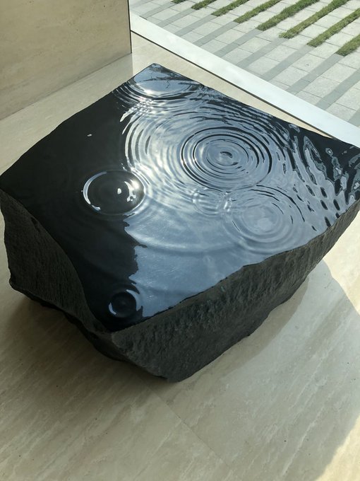 A polished black stone sculpture with a water-like rippled surface creating circular patterns on top, displayed on a wooden floor.