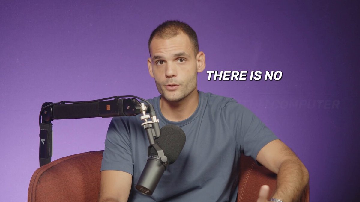 In this image, there's a man sitting inside an audio booth with purple walls behind him. He appears to be giving a speech or discussing a topic on his microphone. The words "There is no" are visible next to him, which could possibly be related to the topic he's talking about or some comment he received while speaking at the event.