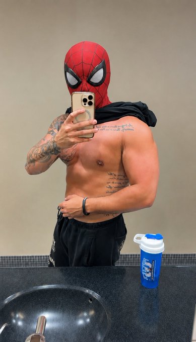 A muscular, shirtless man wearing a Spider-Man mask and black shorts takes a mirror selfie in a bathroom, with a shaker bottle on the counter.