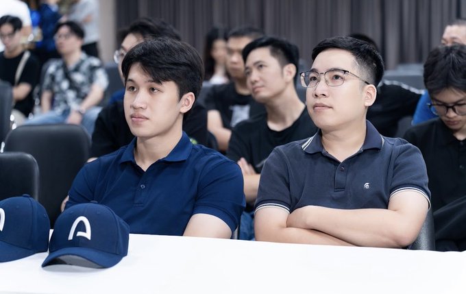 Two men in polo shirts sitting at a table in what appears to be a conference or event setting, with a blue baseball cap featuring a white 'A' logo visible in the foreground.