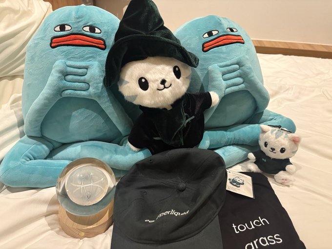 A collection of plushies including two blue creatures, a white bear in a black wizard hat, and a small white cat, displayed with hats and clothing on a bed.