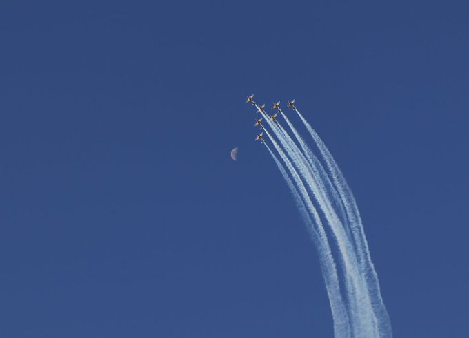 A formation of six small aircraft flies diagonally across a deep blue sky, leaving long white contrails behind them, with a crescent moon visible in the upper portion of the frame.
