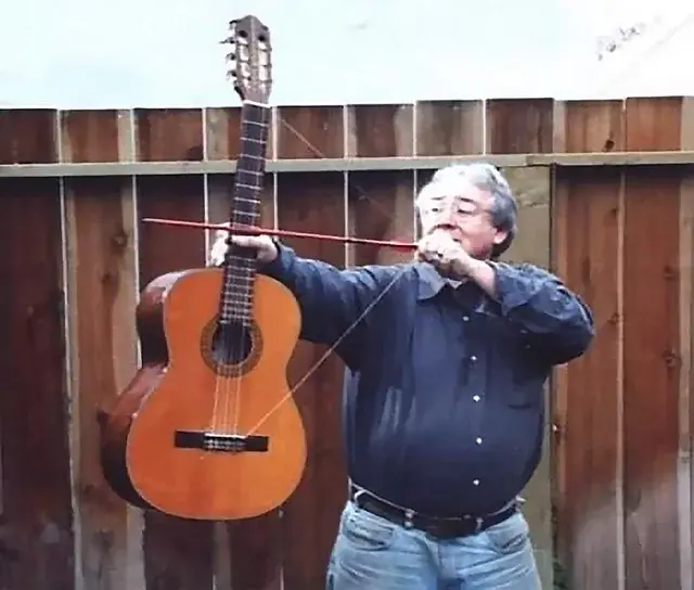 A man wearing glasses and a blue shirt stands in front of a wooden fence, holding a guitar and appearing to play it with a bow like a violin.