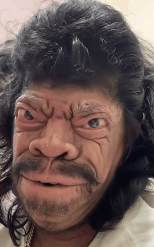 A person wearing a detailed prosthetic mask of a primitive hominid or caveman, featuring a grimacing expression, dark hair, and a beard.