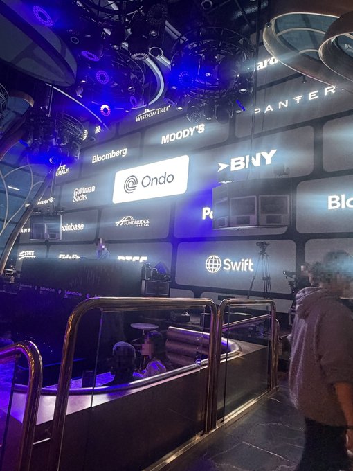 An indoor event space with blue lighting featuring illuminated sponsor logos including Bloomberg, Moody's, Ondo, Binance, Coinbase, and Swift, with attendees visible in the foreground.