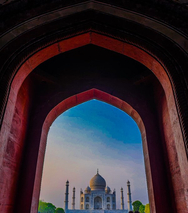The Taj Mahal viewed through a large arched stone doorway with reddish walls, set against a blue and pink sky.