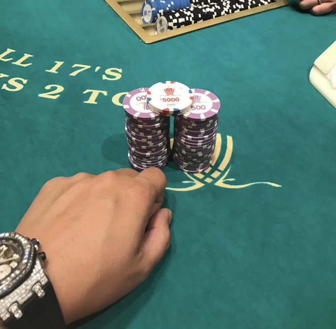 A player's hand rests on a casino table beside a stacked tower of purple $5,000 Casino Aruba chips, with the felt reading "TILL 17'S" and "BETS 2 TO 1," suggesting a high-stakes blackjack game. The post's playful "What we are buying?" caption hints at the allure of stacking serious winnings at the tables.