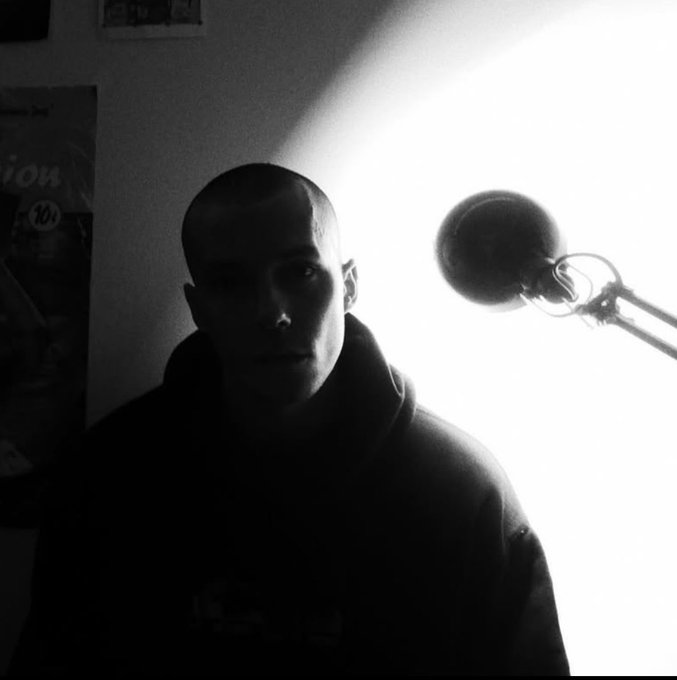 A black and white photo shows a person with a shaved head wearing a hoodie, illuminated by a bright desk lamp in a dimly lit room.
