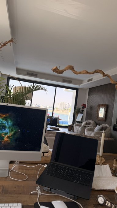 A home office desk setup featuring an iMac with a galaxy wallpaper, an open laptop, a mouse, and a keyboard, with a view of a living room and city skyline through the window.