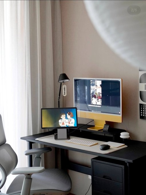 A modern minimalist home office setup featuring a large monitor, a smaller tablet displaying an anime scene, a desk lamp, and an ergonomic chair near a window with white curtains.